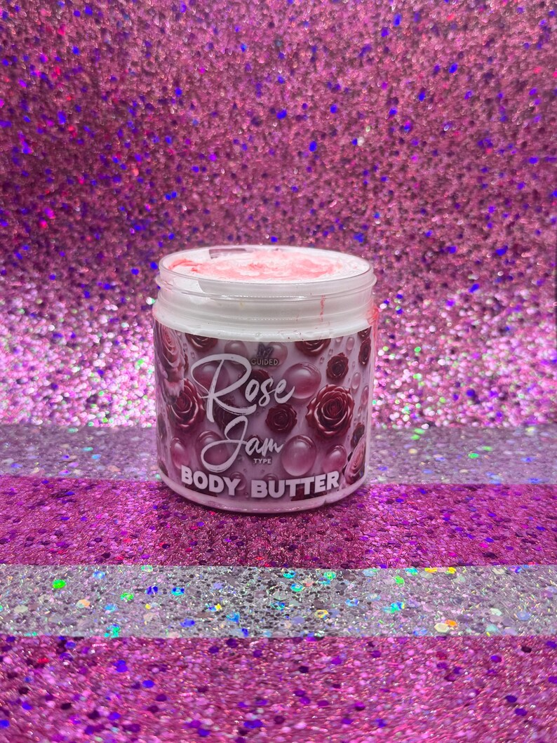 May include: A white jar of body butter with a pink lid. The jar has a label that says "Infused Rose Jam Type Body Butter". The label has a pink and purple floral design.