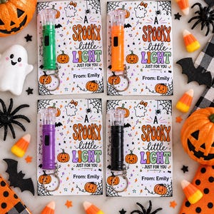May include: Four small plastic flashlights in green, orange, purple, and black, each attached to a metal keychain. They are mounted on white cards featuring Halloween illustrations of ghosts, pumpkins, and webs with the text A SPOOKY little LIGHT JUST FOR YOU From: Emily.