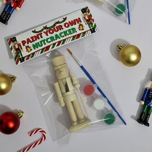 May include: A 'Paint Your Own Nutcracker' kit with a wooden nutcracker figure, paint pots, and a paintbrush. The kit is packaged in a clear plastic bag, with a decorative label. Christmas ornaments and a candy cane are also visible.