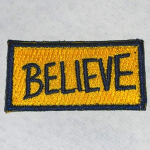 May include: A yellow embroidered patch with a black border. The patch says "BELIEVE" in black embroidery.