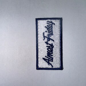 May include: White embroidered patch with blue stitching, featuring the text "Almost Friday" in a cursive font.