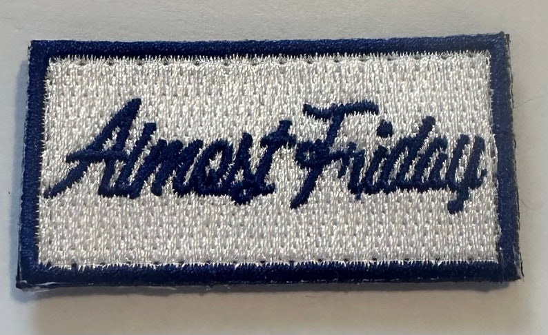 May include: A white embroidered patch with a blue border. The text "Almost Friday" is embroidered in blue cursive lettering.