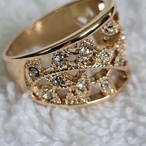 May include: Gold-toned ring with an openwork design. The ring features a series of small, clear gemstones set within the intricate filigree pattern. The band is wide and smooth, creating a contrast with the detailed upper portion.