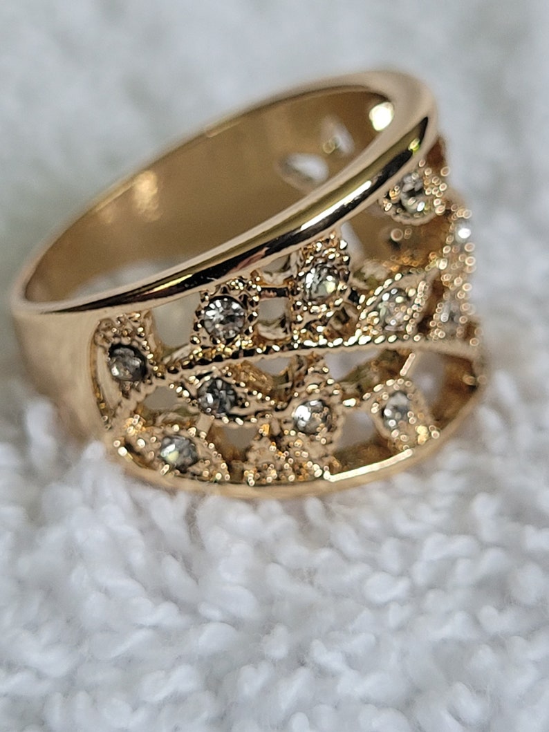 May include: Gold-toned ring with an openwork design. The ring features a series of small, clear gemstones set within the intricate filigree pattern. The band is wide and smooth, creating a contrast with the detailed upper portion.
