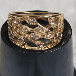 May include: A gold-toned ring with a filigree design featuring leaf-like patterns and small, clear gemstones. The ring has a band and a decorative top, showcasing a delicate and ornate style. The ring is displayed on a black surface.