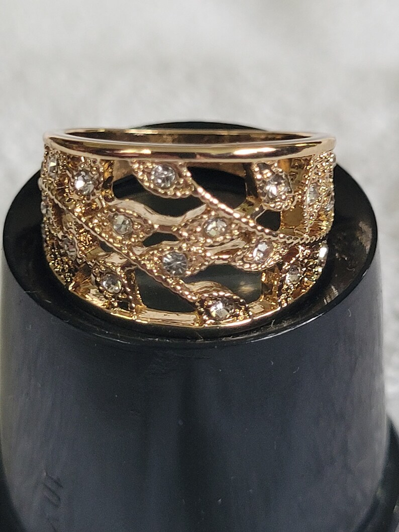 May include: A gold-toned ring with a filigree design featuring leaf-like patterns and small, clear gemstones. The ring has a band and a decorative top, showcasing a delicate and ornate style. The ring is displayed on a black surface.