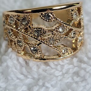 May include: A gold-colored ring with an openwork design. The ring features a series of interconnected, leaf-like shapes, embellished with small, clear gemstones. The ring is set against a white, textured background.