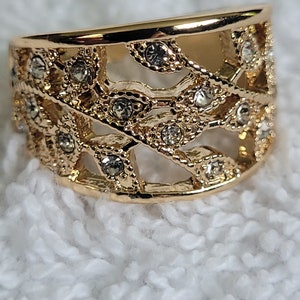May include: A gold-colored ring with an openwork design. The ring features a series of interconnected, leaf-like shapes, embellished with small, clear gemstones. The ring is set against a white, textured background.