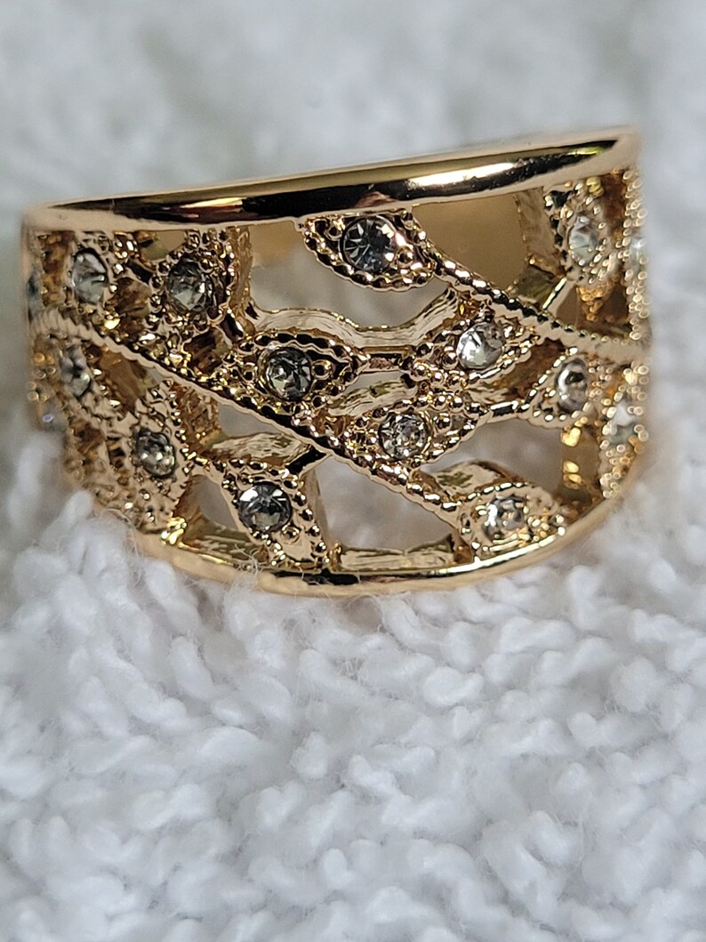 May include: A gold-colored ring with an openwork design. The ring features a series of interconnected, leaf-like shapes, embellished with small, clear gemstones. The ring is set against a white, textured background.