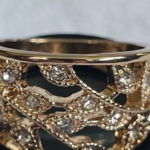 May include: Close-up of a gold-toned ring with an openwork design. The ring features small, clear gemstones set within the intricate pattern. The band has a polished finish, and the overall aesthetic is elegant.