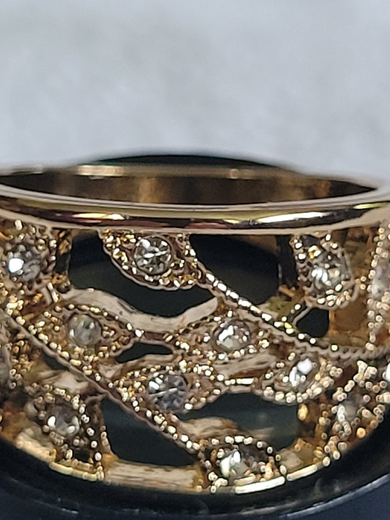 May include: Close-up of a gold-toned ring with an openwork design. The ring features small, clear gemstones set within the intricate pattern. The band has a polished finish, and the overall aesthetic is elegant.