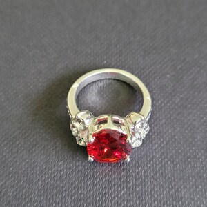 May include: Silver ring featuring a large, oval-cut, red gemstone. The ring's band has skull and crystal accents on each side of the gemstone, creating a gothic or edgy aesthetic. The ring is set against a dark gray background.