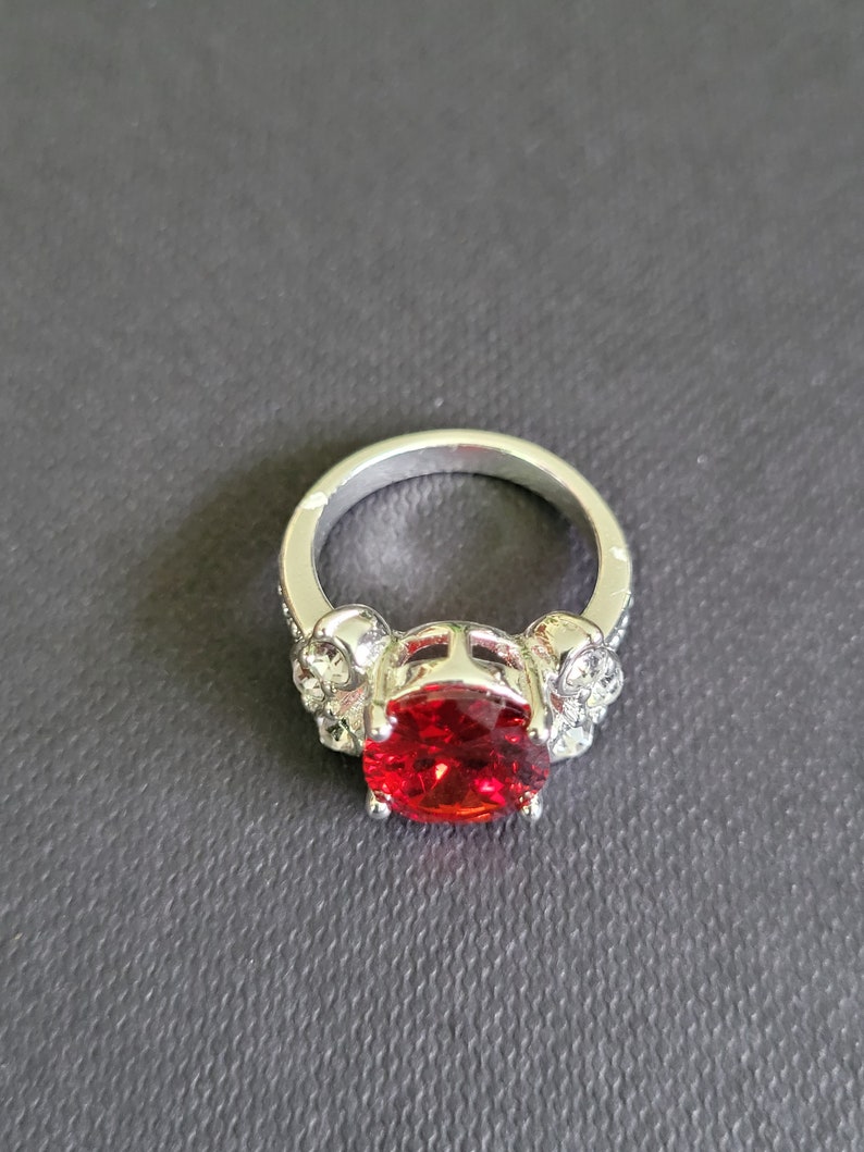 May include: Silver ring featuring a large, oval-cut, red gemstone. The ring's band has skull and crystal accents on each side of the gemstone, creating a gothic or edgy aesthetic. The ring is set against a dark gray background.