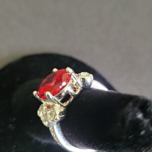 May include: A silver ring featuring a large, marquise-cut, red gemstone as the centerpiece. The ring has a decorative setting with smaller, clear stones on the sides. The ring is displayed on a black surface.