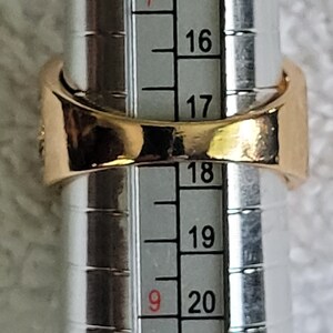 May include: A gold-colored ring is being measured on a ring sizer. The ring is a simple band design. The ring sizer shows a size of approximately 19, which is equivalent to a ring size of 9.
