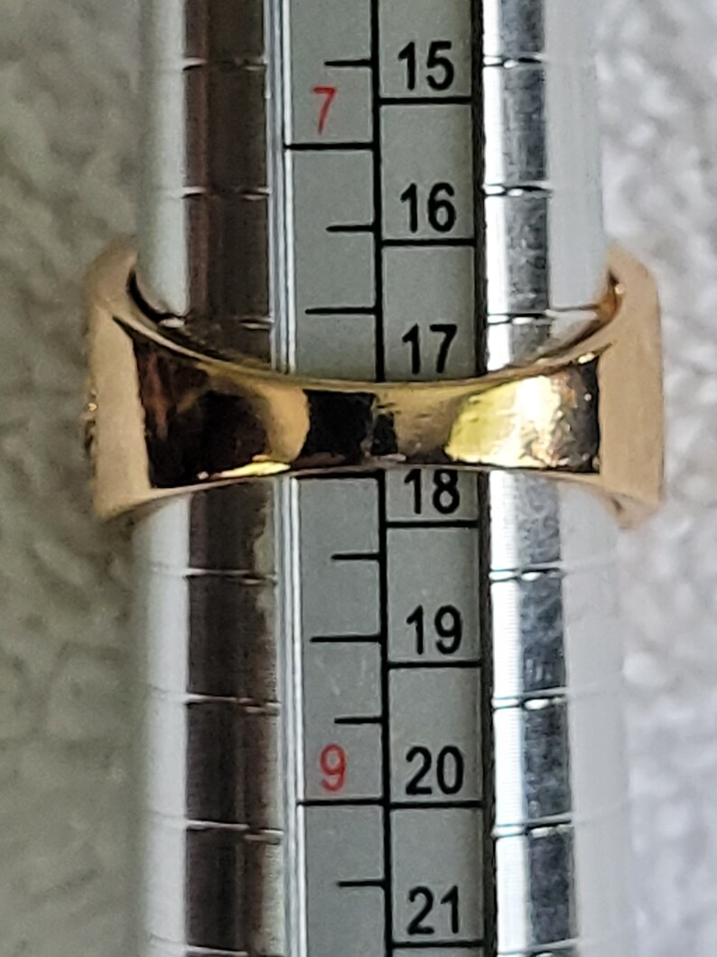 May include: A gold-colored ring is being measured on a ring sizer. The ring is a simple band design. The ring sizer shows a size of approximately 19, which is equivalent to a ring size of 9.