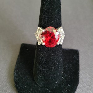 May include: A silver ring with a large, oval-cut, bright red gemstone as the centerpiece. The ring has a decorative design with smaller, clear stones on either side of the main stone. The ring is displayed on a black ring holder.