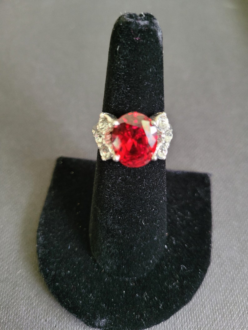May include: A silver ring with a large, oval-cut, bright red gemstone as the centerpiece. The ring has a decorative design with smaller, clear stones on either side of the main stone. The ring is displayed on a black ring holder.