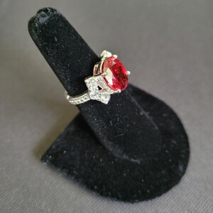 May include: A silver-toned ring with a large, oval-shaped, red gemstone as the centerpiece. The ring is adorned with smaller, clear gemstones and is displayed on a black ring holder against a gray background. The ring's design is intricate and detailed.