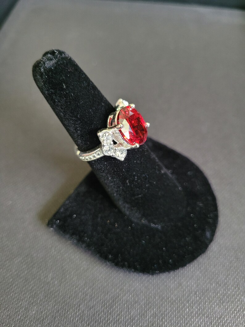 May include: A silver-toned ring with a large, oval-shaped, red gemstone as the centerpiece. The ring is adorned with smaller, clear gemstones and is displayed on a black ring holder against a gray background. The ring's design is intricate and detailed.