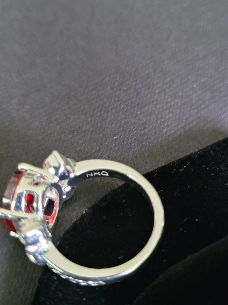 May include: A silver ring with a large, oval-shaped, red gemstone as the centerpiece. The ring has a detailed setting with smaller accent stones and the inscription "NRQ" on the band. The ring is set against a dark background.