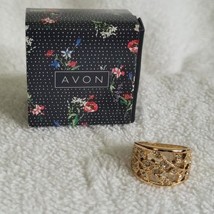 May include: A gold-toned ring with a decorative openwork design and small clear stones. The ring sits next to a black box with a floral pattern and the word "AVON" in white.