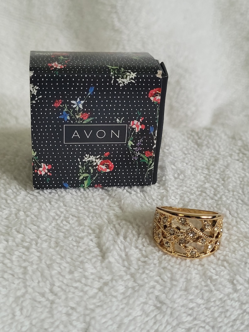 May include: A gold-toned ring with a decorative openwork design and small clear stones. The ring sits next to a black box with a floral pattern and the word "AVON" in white.