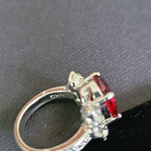May include: A silver ring featuring a large, oval-cut, deep red gemstone as the centerpiece. The ring is accented with smaller, clear gemstones and has the inscription "NRQ" on the band.