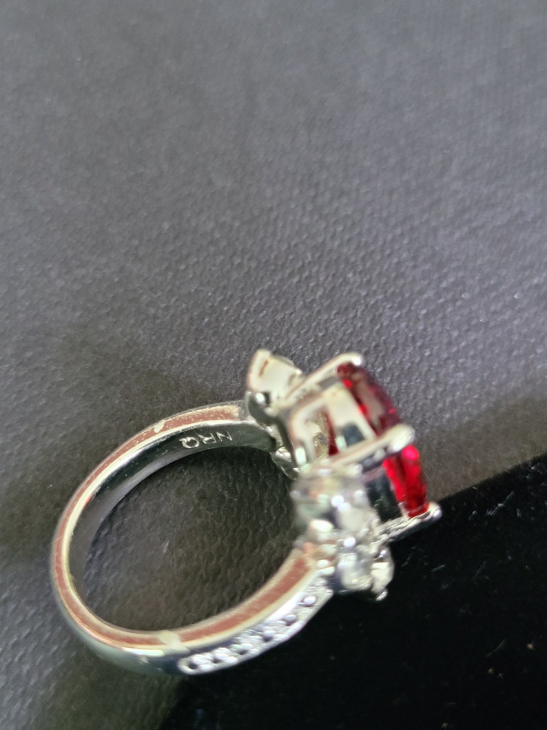 May include: A silver ring featuring a large, oval-cut, deep red gemstone as the centerpiece. The ring is accented with smaller, clear gemstones and has the inscription "NRQ" on the band.
