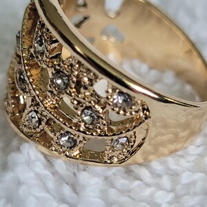 May include: Close-up of a gold-colored ring with a decorative design. The ring features a series of small, clear gemstones set within an openwork pattern. The band has a smooth, polished finish, while the decorative section has a textured appearance.