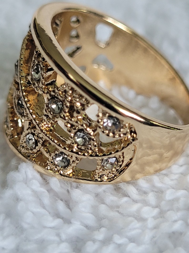 May include: Close-up of a gold-colored ring with a decorative design. The ring features a series of small, clear gemstones set within an openwork pattern. The band has a smooth, polished finish, while the decorative section has a textured appearance.