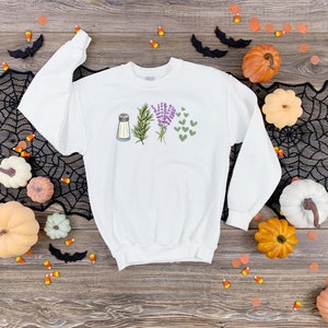 May include: A white long-sleeve sweatshirt featuring a graphic print on the chest. The design displays a silver salt shaker, a green sprig of rosemary, a bundle of purple lavender flowers, and a cluster of small green heart shapes arranged in a row.