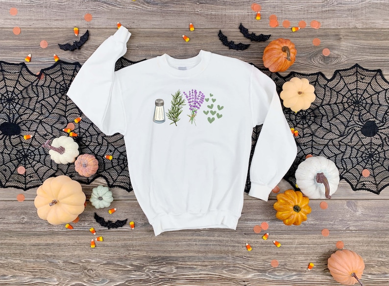 May include: A white long-sleeve sweatshirt featuring a graphic print on the chest. The design displays a silver salt shaker, a green sprig of rosemary, a bundle of purple lavender flowers, and a cluster of small green heart shapes arranged in a row.