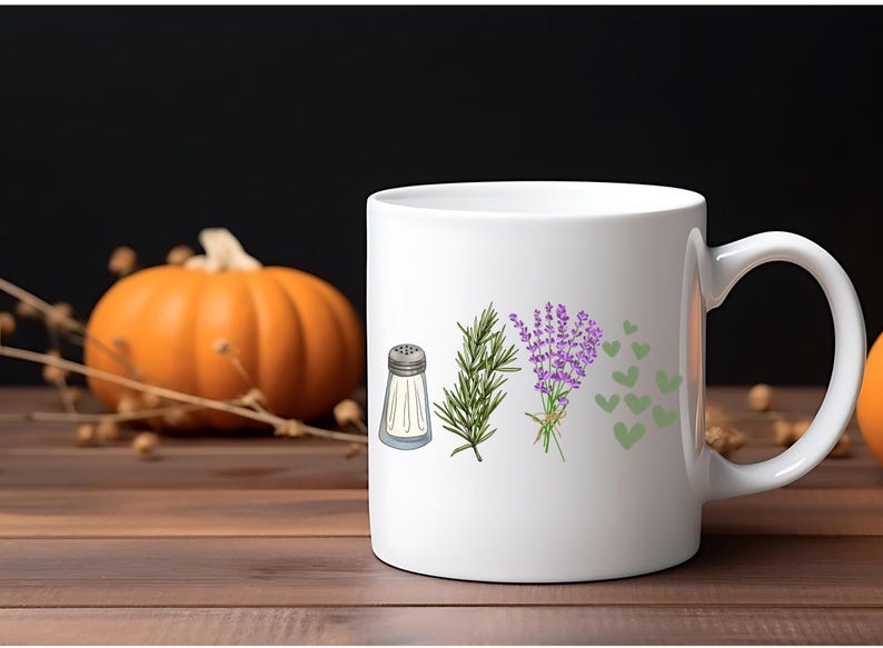 May include: A white ceramic mug featuring a printed graphic of a silver salt shaker, a sprig of green rosemary, a bundle of purple lavender, and several small green hearts arranged in a horizontal row on the front surface of the mug.