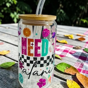 May include: A clear glass tumbler with a bamboo lid and a clear straw. The tumbler is decorated with the words "NEED safari" in colorful letters and various tropical-themed graphics, including a flamingo and flowers.