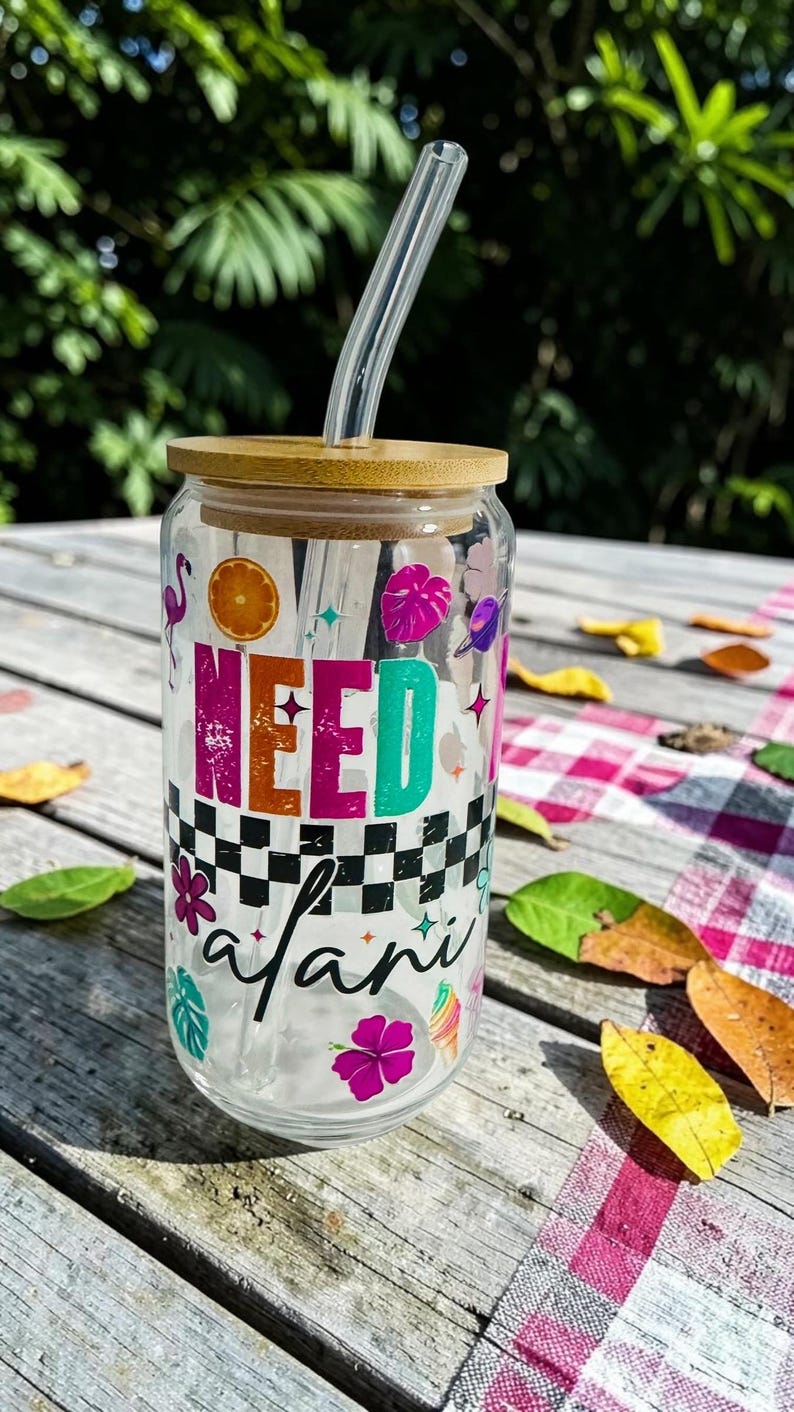 May include: A clear glass tumbler with a bamboo lid and a clear straw. The tumbler is decorated with the words "NEED safari" in colorful letters and various tropical-themed graphics, including a flamingo and flowers.