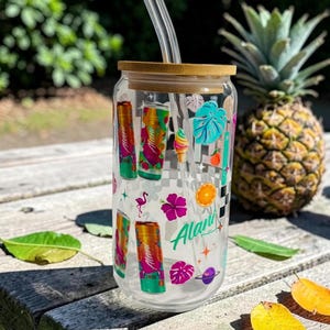 May include: A clear glass tumbler with a bamboo lid and a clear straw. The tumbler is decorated with colorful tropical-themed graphics, including cans, flowers, and fruit. A pineapple sits in the background.