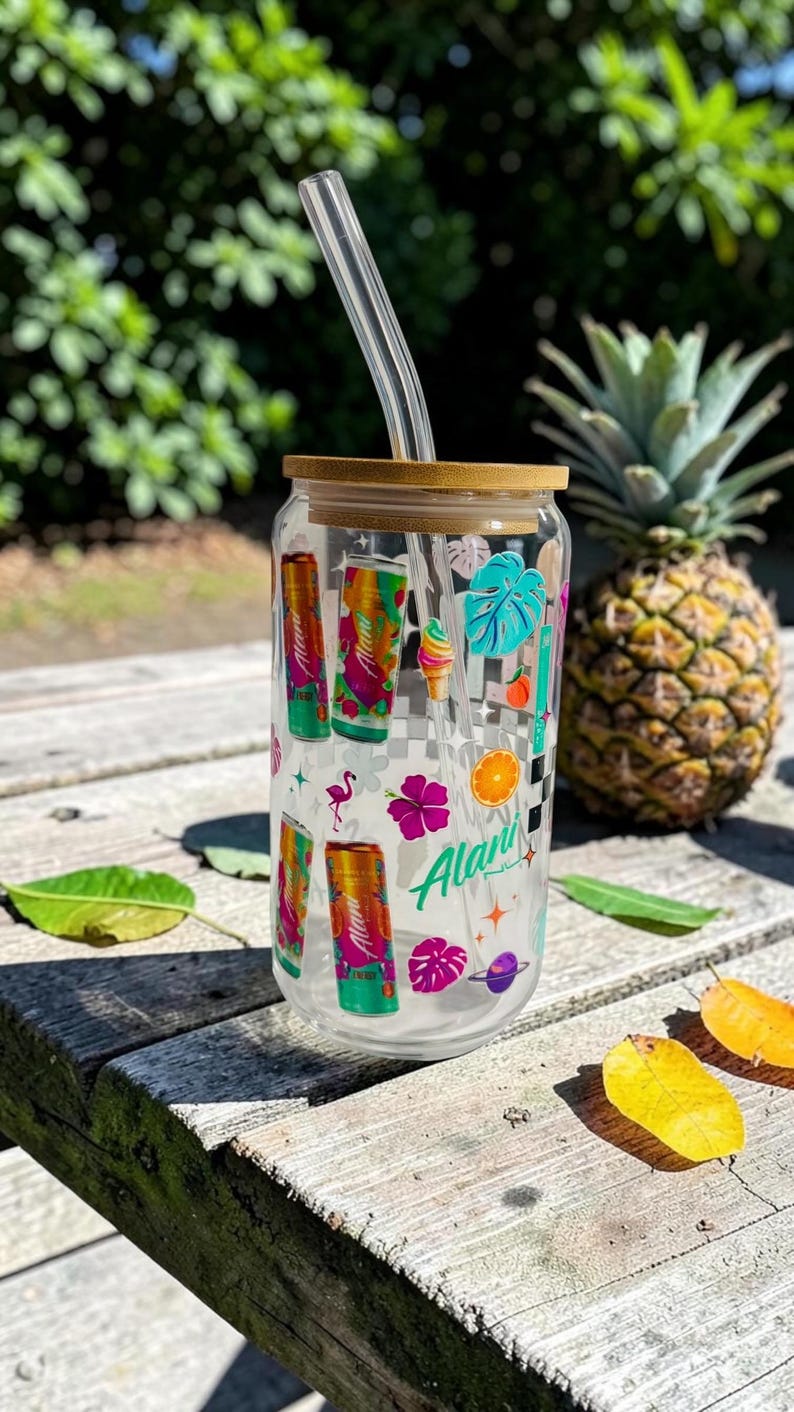 May include: A clear glass tumbler with a bamboo lid and a clear straw. The tumbler is decorated with colorful tropical-themed graphics, including cans, flowers, and fruit. A pineapple sits in the background.