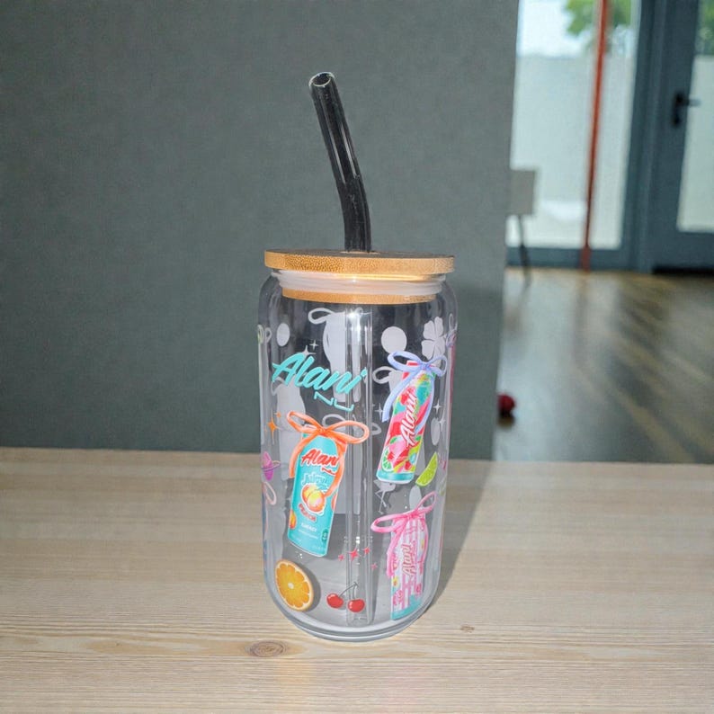 May include: A clear glass tumbler with a bamboo lid and a black straw. The tumbler is decorated with colorful graphics of canned beverages, fruit slices, and the text "Alan".