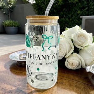 May include: A clear glass tumbler with a bamboo lid and a glass straw. The tumbler is decorated with green and black graphics and the text "TIFFANY & CO. NEW YORK SINCE 1837". A silver bracelet is visible inside the tumbler.