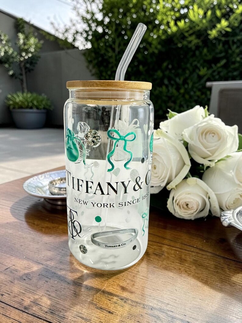 May include: A clear glass tumbler with a bamboo lid and a glass straw. The tumbler is decorated with green and black graphics and the text "TIFFANY & CO. NEW YORK SINCE 1837". A silver bracelet is visible inside the tumbler.