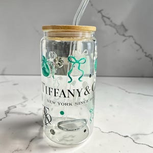 May include: A clear glass tumbler with a wooden lid and a glass straw. The tumbler is decorated with green and silver graphics, including gift bags and bows. The text "TIFFANY & CO. NEW YORK SINCE 1837" is printed on the front.