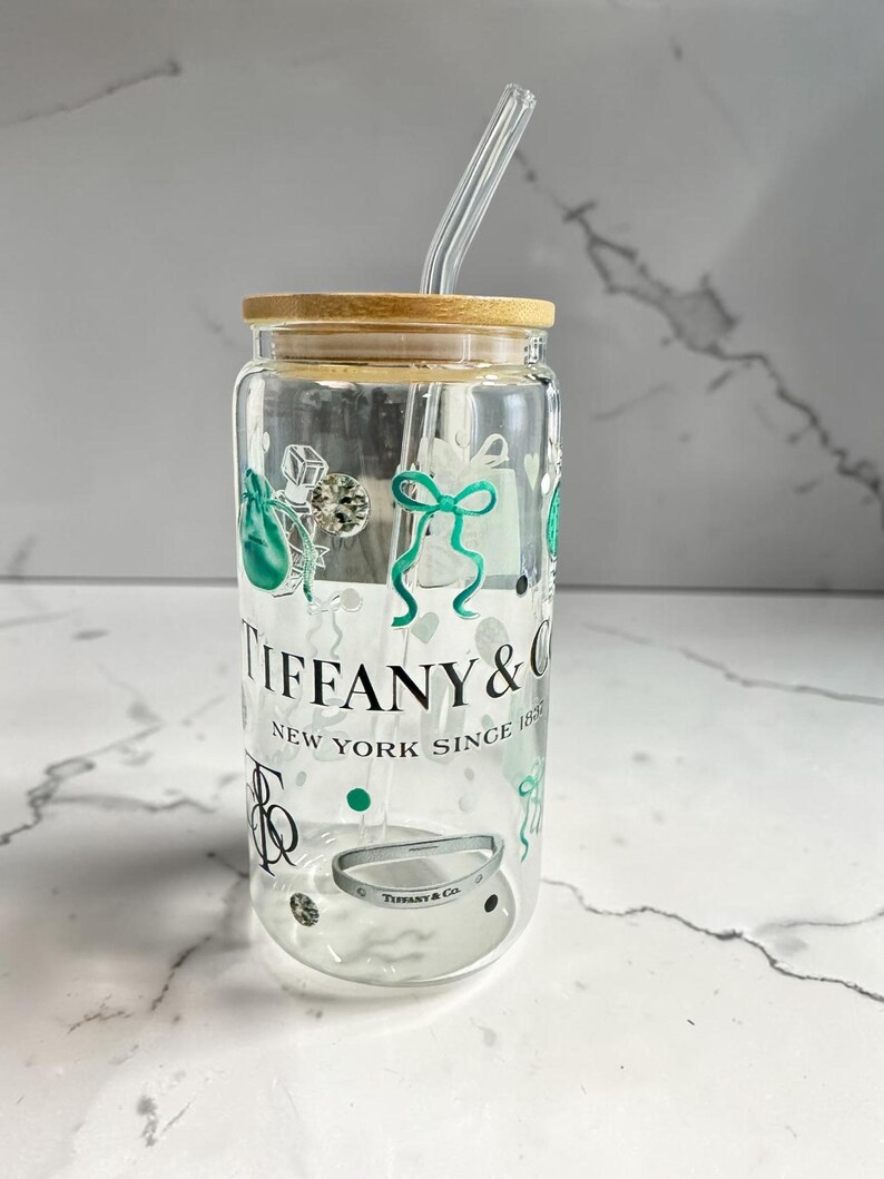May include: A clear glass tumbler with a wooden lid and a glass straw. The tumbler is decorated with green and silver graphics, including gift bags and bows. The text "TIFFANY & CO. NEW YORK SINCE 1837" is printed on the front.