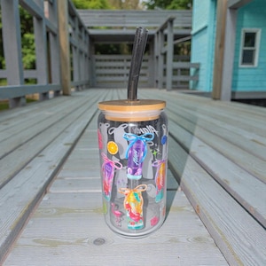 May include: A clear glass tumbler with a bamboo lid and a black straw. The tumbler is decorated with colorful graphics of tropical drinks, a flamingo, and the word "Aloha". The tumbler is sitting on a wooden deck.