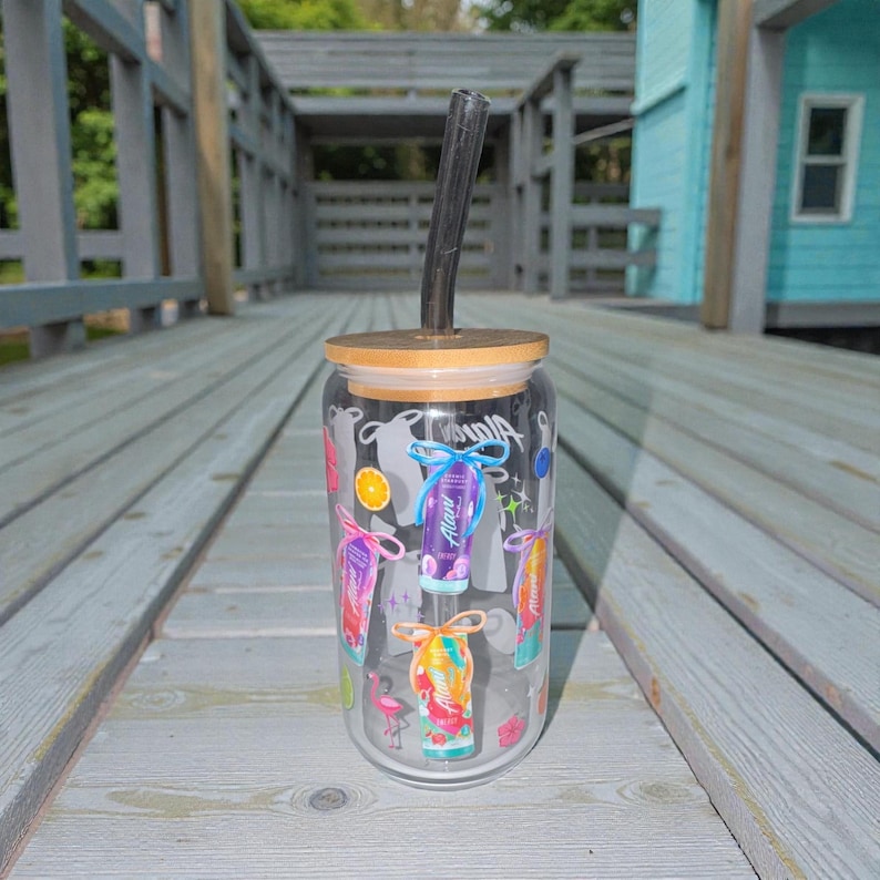 May include: A clear glass tumbler with a bamboo lid and a black straw. The tumbler is decorated with colorful graphics of tropical drinks, a flamingo, and the word "Aloha". The tumbler is sitting on a wooden deck.