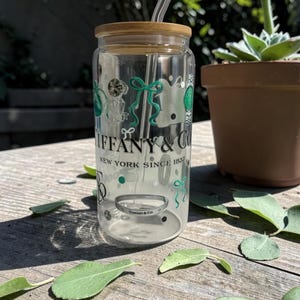 May include: A clear glass tumbler with a bamboo lid and a clear straw. The tumbler features the "Tiffany & Co." logo and decorative green and white graphics. The text "NEW YORK SINCE 1837" is also visible.