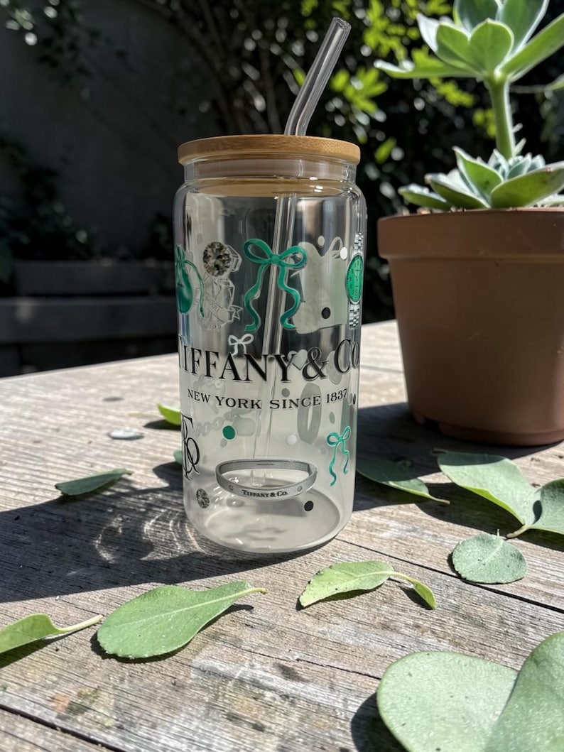 May include: A clear glass tumbler with a bamboo lid and a clear straw. The tumbler features the "Tiffany & Co." logo and decorative green and white graphics. The text "NEW YORK SINCE 1837" is also visible.