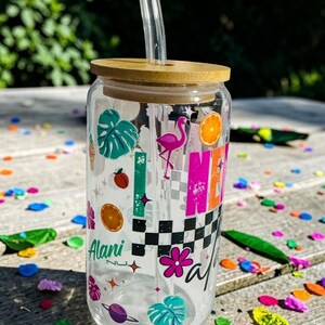 May include: A clear glass tumbler with a bamboo lid and a clear straw. The tumbler is decorated with colorful graphics including a flamingo, orange slices, and the name "Alani". The design has a checkered pattern and tropical leaves.