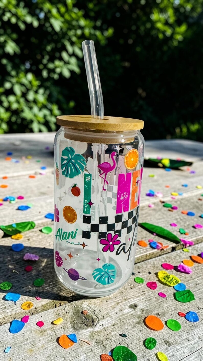 May include: A clear glass tumbler with a bamboo lid and a clear straw. The tumbler is decorated with colorful graphics including a flamingo, orange slices, and the name "Alani". The design has a checkered pattern and tropical leaves.