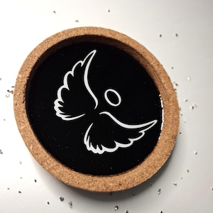 May include: Round coaster with a black center featuring a white angel design. The coaster has a natural cork border. The angel graphic has outstretched wings and a simple halo. Decorative item for home decor.